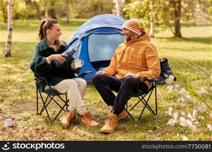 camping, tourism and travel concept - happy couple with thermos pouring tea to cup at tent camp. happy couple with thermos pouring tea at tent camp