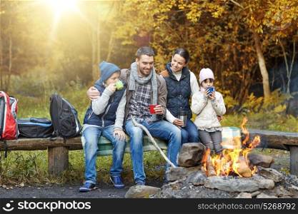 camping, travel, tourism and people concept - happy family sitting on bench and drinking hot tea from cups at camp fire in autumn forest. happy family sitting on bench at camp fire