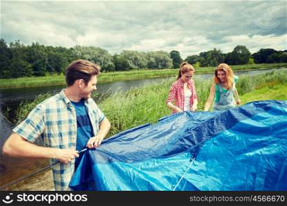 camping, travel, tourism, hike and people concept - group of smiling friends setting up tent outdoors