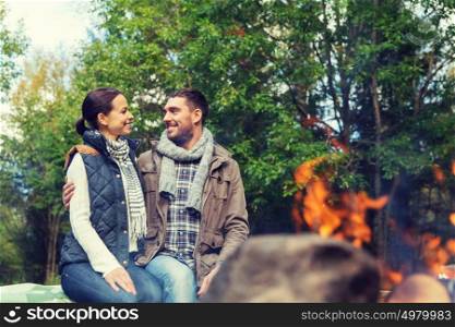 camping, travel, tourism, hike and people concept - happy couple sitting on bench and warming near campfire at camp in woods. happy couple sitting on bench near camp fire