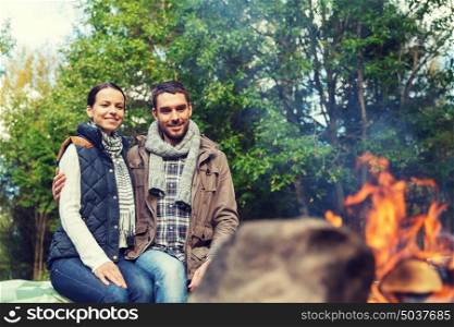 camping, travel, tourism, hike and people concept - happy couple sitting on bench and warming near campfire at camp in woods. happy couple sitting on bench near camp fire