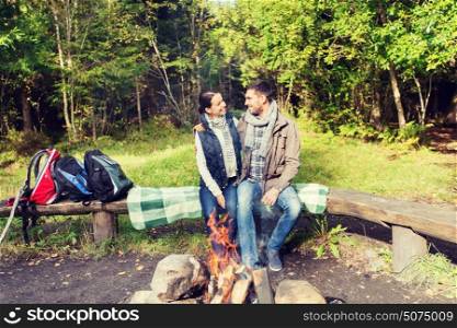 camping, travel, tourism, hike and people concept - happy couple sitting on bench and hugging near campfire at camp in woods. happy couple sitting on bench near camp fire