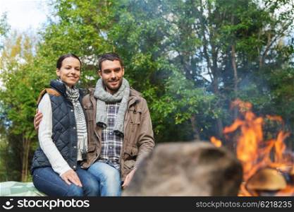 camping, travel, tourism, hike and people concept - happy couple sitting on bench and warming near campfire at camp in woods
