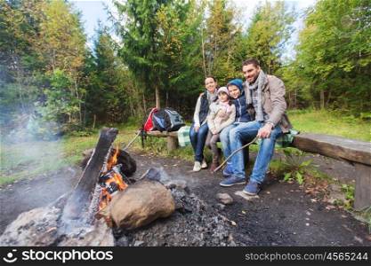 camping, travel, tourism, hike and people concept - happy family sitting on bench at camp fire in woods