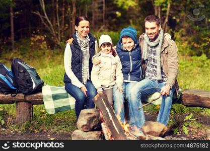 camping, travel, tourism, hike and people concept - happy family sitting on bench at camp fire in woods
