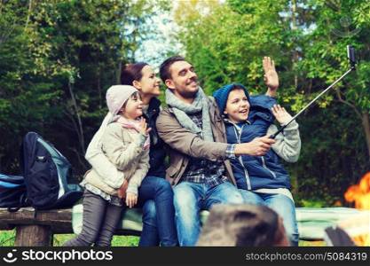 camping, travel, tourism, hike and people concept - happy family sitting on bench and taking picture with smartphone on selfie stick at campfire in woods. family with smartphone taking selfie near campfire. family with smartphone taking selfie near campfire