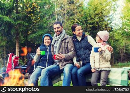 camping, travel, tourism, hike and people concept - happy family sitting on bench and drinking hot tea from cups at camp fire in woods. happy family sitting on bench at camp fire. happy family sitting on bench at camp fire