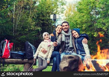 camping, travel, tourism, hike and people concept - happy family sitting on bench and taking picture with smartphone on selfie stick at campfire in woods. family with smartphone taking selfie near campfire