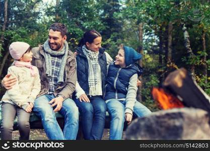 camping, travel, tourism, hike and people concept - happy family sitting on bench and talking at camp near campfire in woods. happy family sitting on bench at camp fire