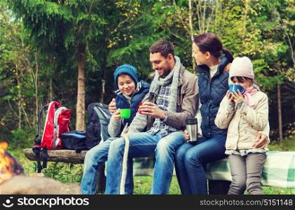camping, travel, tourism, hike and people concept - happy family sitting on bench and drinking hot tea from cups at camp fire in woods. happy family sitting on bench at camp fire