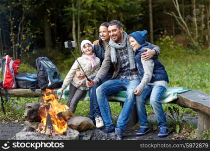 camping, travel, tourism, hike and people concept - happy family sitting on bench and taking picture with smartphone on selfie stick at campfire in woods