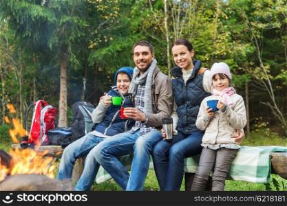 camping, travel, tourism, hike and people concept - happy family sitting on bench and drinking hot tea from cups at camp fire in woods