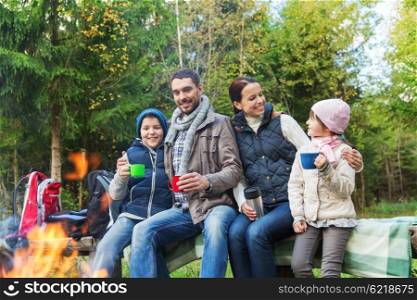 camping, travel, tourism, hike and people concept - happy family sitting on bench and drinking hot tea from cups at camp fire in woods