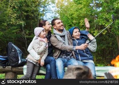 camping, travel, tourism, hike and people concept - happy family sitting on bench and taking picture with smartphone on selfie stick at campfire in woods