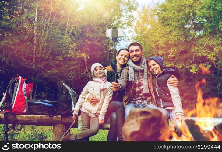 camping, travel, tourism, hike and people concept - happy family sitting on bench and taking picture with smartphone on selfie stick at campfire in woods. family with smartphone taking selfie near campfire