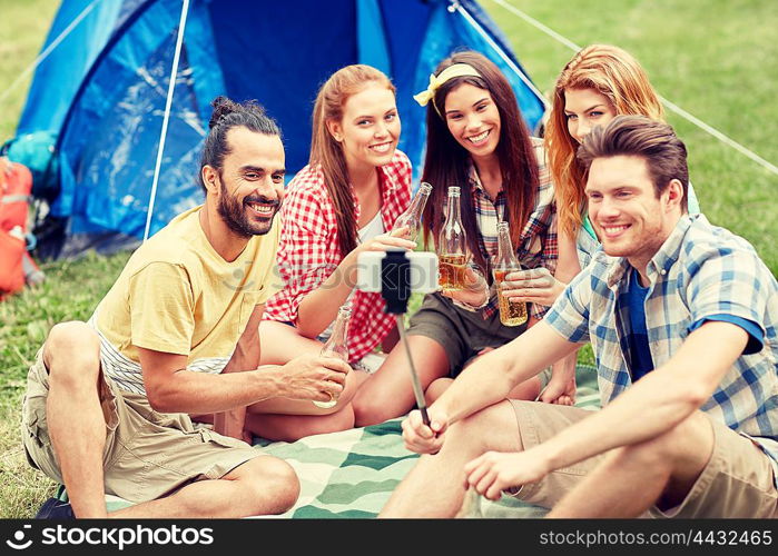 camping, travel, tourism, hike and people concept - happy friends with glass bottles drinking cider or beer and taking picture by smartphone on selfie stick at camping