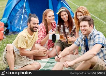 camping, travel, tourism, hike and people concept - happy friends with glass bottles drinking cider or beer and taking picture by smartphone on selfie stick at camping