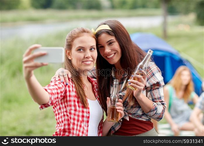 camping, travel, tourism, hike and people concept - happy young women with glass bottles drinking cider or beer and taking selfie by smartphone at camping