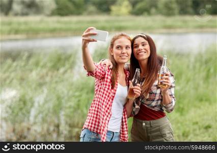 camping, travel, tourism, hike and people concept - happy young women with glass bottles drinking cider or beer and taking selfie by smartphone outdoors