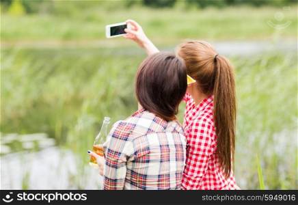 camping, travel, tourism, hike and people concept - happy young women with glass bottles drinking cider or beer and taking selfie by smartphone outdoors from back