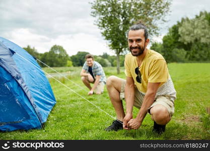 camping, travel, tourism, hike and people concept - smiling male friends setting up tent outdoors