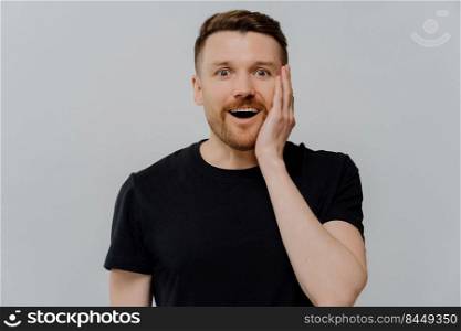 Can not believe its true. Young handsome ginger man in black tshirt dropping jaw from excitement, being amazed shocked with something unbelievable while standing against grey wall. Amazed ginger man staring at camera with shocked happy expression