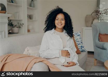 Can not wait to become mom. Positive smiling p®nant afro american woman in knitted bei≥dress touχng≥ntly her belly and looking with smi≤aside at home. P®nancy and expectation concept. Happy afro american p®nant fema≤happy to become mom in future