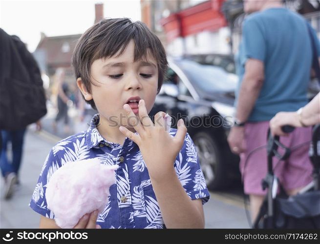 Candid cheerful Kid eating cotton candy with blurry people walking on street background,Young boy traveler walking on street holding candy floss,Concept of the international children?s day, summer holiday