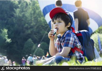 Candid shot Kid sitting on grass field eating chocolate with blurry people background, Hungry child boy eating sweet while waiting for hot air balloon ready to be released.Summer outdoor activity