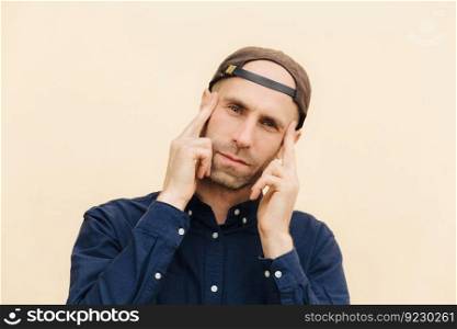 Candid shot of attractive unshaven male looks thoughtfully and concetrated at camera, keeps fore fingers on temples, wears stylish cap and shirt, isolated over light background. People and style