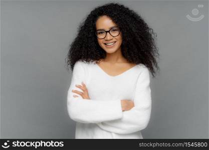 Candid shot of beautiful African American woman with curls, has gentle smile, keeps hands crossed, wears white soft sweater, stands against grey background. Positive emotions and feelings concept