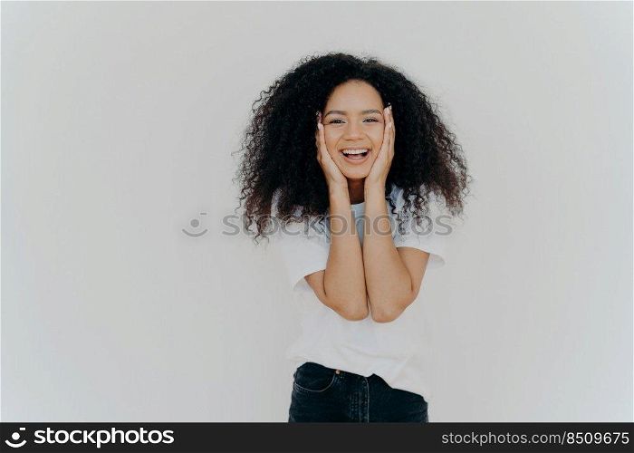 Candid shot of good looking lady with curly hairstyle, smiles cute, keeps both hands on cheeks, shows positive emotions, wears white t shirt, black denim trousers, stands indoor, blank copy space