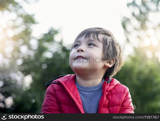 Candid shot of happy little kid looking up with smiling face sitting in the park,Cropped shot of child boy looking up to sky relaxing on his own with blurry natural green forest backgroud, Positive children concept