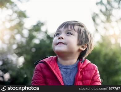 Candid shot of happy little kid looking up with smiling face sitting in the park,Cropped shot of child boy looking up to sky relaxing on his own with blurry natural green forest backgroud, Positive children concept