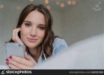 Candid shot of pretty dark haired woman spneds free time with favourite song, holds modern cell phone, looks directly at camera, poses indoor, has make up. Attractive girl searches new melodies