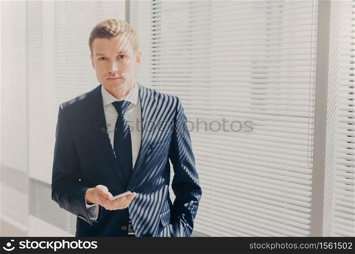 Candid shot of prosperous businessman checks notification on cellular, plans daily schedule, wears formal suit, stands near window, looks directly into camera, has work break. Office routine