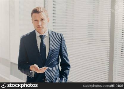Candid shot of prosperous businessman checks notification on cellular, plans daily schedule, wears formal suit, stands near window, looks directly into camera, has work break. Office routine