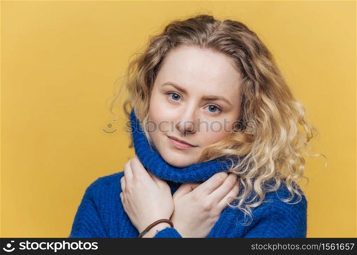 Candid shot of serious adorable woman with curly hair, wears warm blue sweater, warms herself after walk outdoor, isolated over yellow studio background. Attractive female has confident expression