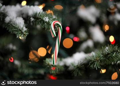 Candy cane ornament hanging in artificial Christmas tree with glowing lights and snow in background
