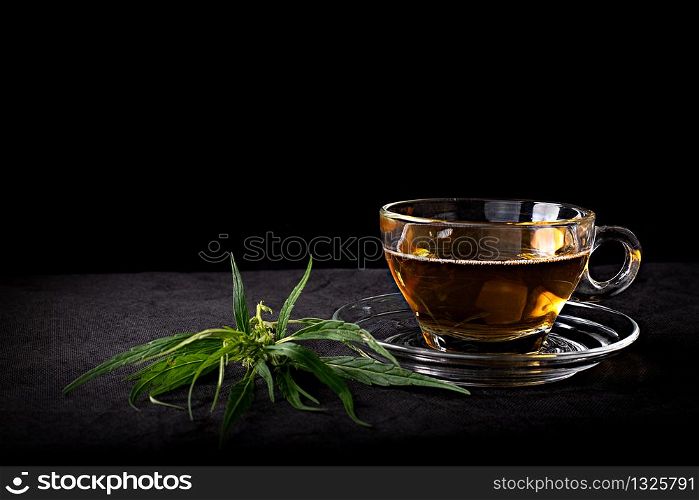 Cannabis tea in a clear glass cup with a green marijuana leaf and stem on wood table at the black background, Herbal and healthy drinks concept