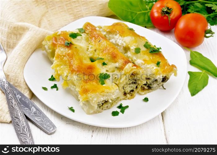 Cannelloni stuffed with cottage cheese and spinach with Bechamel sauce in a plate, towel, tomatoes and parsley on the background of light wooden board