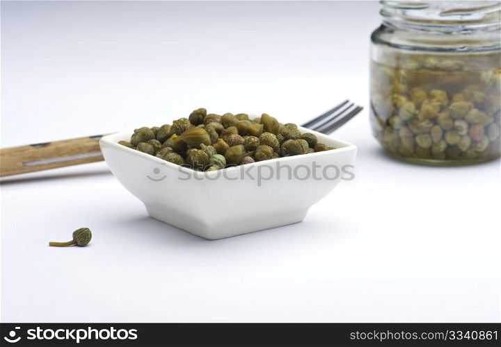 Capers In A White Dish, Against A White Background, With A Jar Of Capers and A Fork In The Background