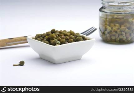 Capers In A White Dish, Against A White Background, With A Jar Of Capers and A Fork In The Background