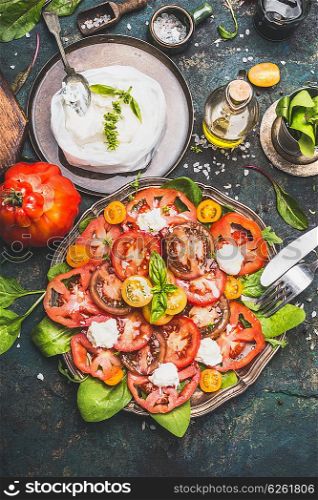 Caprese tomatoes mozzarella salad in aged metal plate with homemade chees and ingredients on dark rustic background, top view