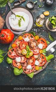 Caprese tomatoes mozzarella salad in aged metal plate with homemade chees and ingredients on dark rustic background, top view