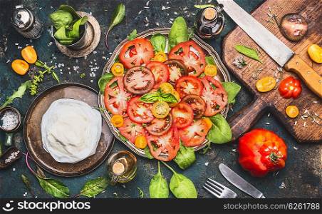 Caprese tomatoes mozzarella salad on dark rustic background with oil, balsamic vinegar, cutting board and ingredients, top view