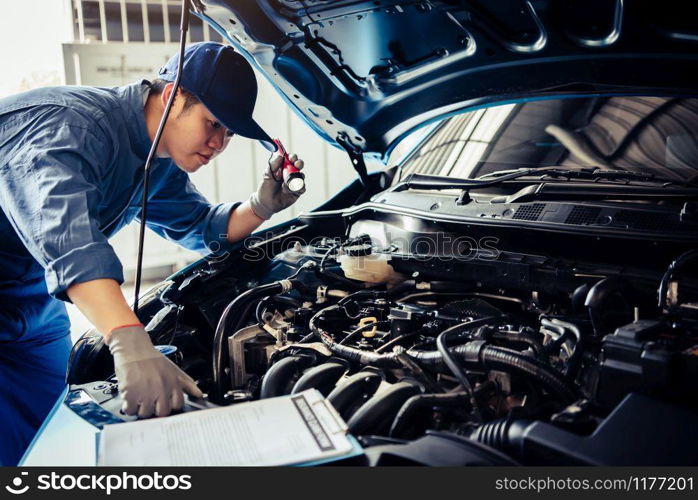 Car mechanic technician holding flashlight checking engine with checklist clipboard to maintenance vehicle by customer claim order in auto repair shop garage repair service. People occupation business