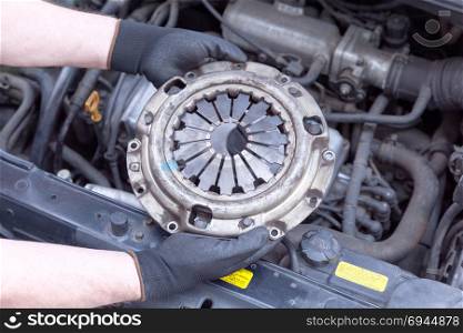 Car mechanic wearing protective work gloves holds old clutch basket over a car engine
