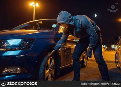 Car robber punctures the tyre, vandalism, hooligan. Hooded male bandit spoils vehicle on parking. Auto robbery, automobile crime