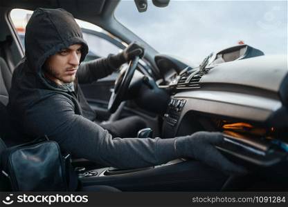 Car robber searches the glove compartment, dangerous hobby, stealing. Hooded male bandit opening vehicle on parking. Auto robbery, automobile crime